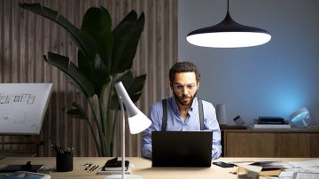 Un hombre se sienta en un escritorio con una computadora portátil frente a él, iluminado por una luz suspendida desde arriba y una lámpara de mesa a su derecha