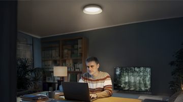 Un homme est assis à un bureau avec un ordinateur portable, éclairé par un plafonnier rond.