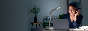 A woman is talking on the phone while she works in her home office, with a small table lamp beside her