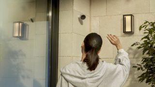 Een vrouw bij een voordeur zwaait naar een Hue Secure beveiligingscamera die aan de muur is bevestigd. 