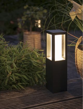 Een zwarte slimme sokkellamp in een tuin, stralend met warm wit licht.