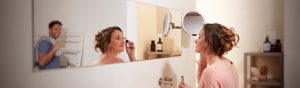 Person applying makeup at a bathroom mirror with bright lighting supporting detailed grooming, bottles and shelves in the background