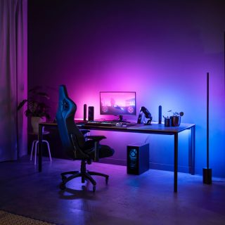 Gaming room setup with smart lighting casting purple and blue hues on wall behind desk, monitor, and vertical floor lamp.