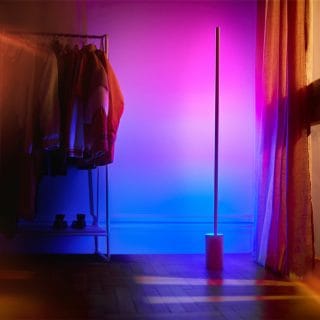 Vertical smart floor lamp placed near a curtain, casting gradient blue and pink light onto the wall and floor.