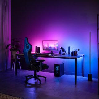 Gaming desk setup with smart lighting devices placed behind monitor and beside desk, casting purple and blue light onto wall and surface.