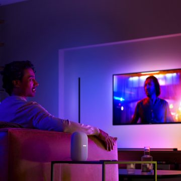 Un hombre en un sofá viendo la televisión con iluminación envolvente y un altavoz inteligente en la mesa auxiliar junto a él