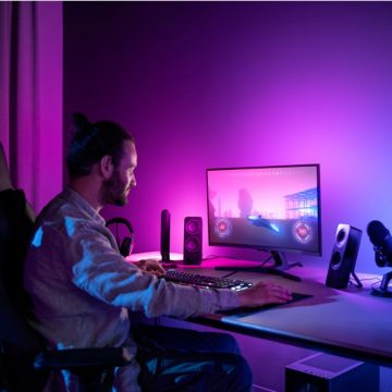 Person gaming at a desk with a computer monitor, surrounded by speakers and accessories, illuminated by vibrant purple lighting.