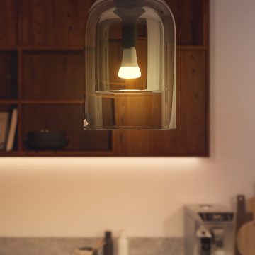 A smart light bulb emitting white light is installed in a glass pendant fixture above a kitchen counter with wooden shelves.