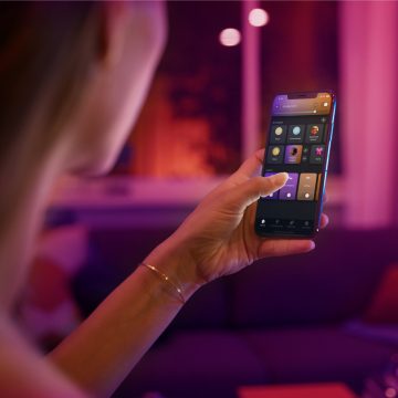 Person using a smartphone app to control smart lighting in a living room with colorful ambient lights in the background.