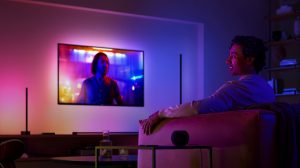 Person seated on a sofa in a living room watching TV, with colored smart lighting illuminating the wall behind the screen.