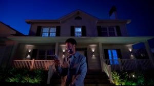 Person using a smartphone on the porch of a house at night, with outdoor smart lights illuminating the entrance and garden.