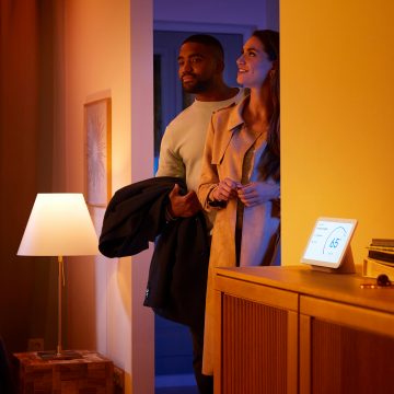 Two people enter a warmly lit home, illuminated by a table lamp and a smart display showing a temperature setting in the hallway.