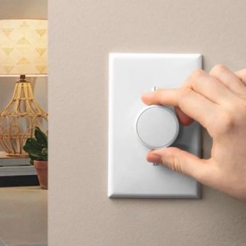 Hand rotating a round white wall dimmer switch to adjust lighting, with a lamp and plant visible in the background.