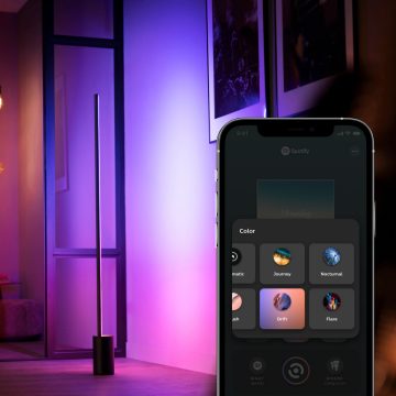 A vertical LED light panel placed near a wall emits purple and pink hues, with a smartphone displaying color control options.