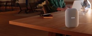 Person working at a desk with a smart speaker in focus, illuminated by warm ambient lighting in a modern living room setting.