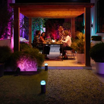 A group of people sharing a meal at an outdoor table under a pergola, surrounded by colorful smart garden lighting.
