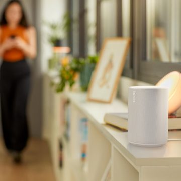 A white smart speaker on a shelf in a modern living room, with ambient colored lighting illuminating nearby décor and books.