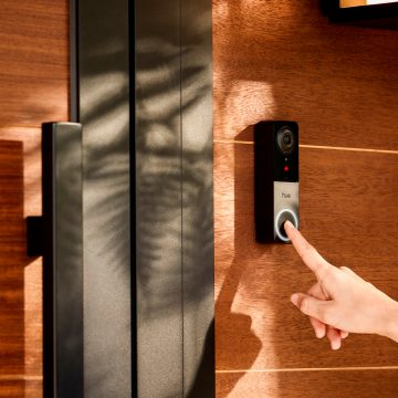 Finger pressing the button on a black smart video doorbell mounted on a wooden wall near a metal door handle.