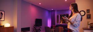 A person using a smartphone to adjust smart lighting in a modern living room with colored lights on ceiling and side table.