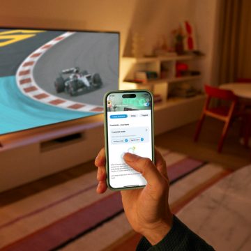 Een hand die een telefoon vasthoudt en de WiZ app gebruikt. Een tv die op de achtergrond een race toont, verlicht door slimme WiZ verlichting.