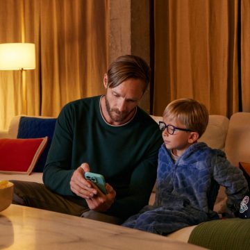 A father showing his phone to his young son on the couch. A lamp with a WiZ Smart Bulb casts warm light.