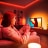 Eine Frau spielt auf ihrem TV mit einer immersiven Entertainment-Beleuchtung in rotem, orangefarbenen und weißem smarten Licht. 
