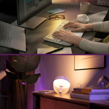 Person tippt an Tastatur mit Notizblock, Tasse und Bildschirm; darunter warme Tischlampe auf Sideboard mit Büchern und Brille.