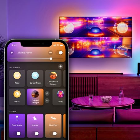 A colorfully lit room with a wall-mounted TV displaying a split-screen racing game. An overlay of the Hue app displays the Home tab.