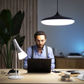 A man sits at a laptop on a desk lit with a smart table lamp and pendant light set to cool white tones.