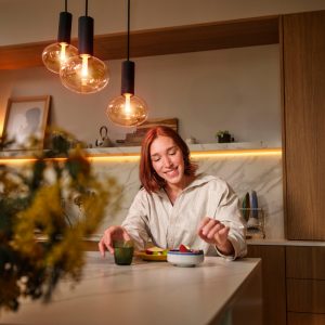 A person sits at a kitchen counter with warm pendant and under-cabinet lighting, enjoying a meal and a drink.