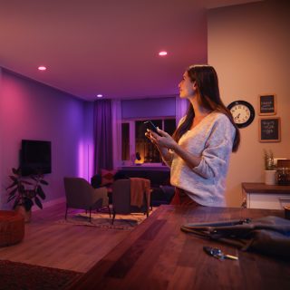 Person adjusting smart ambient lighting in a modern living room using a mobile device, with colored lights illuminating seating area.