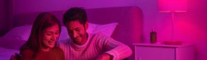 Couple sitting together on a bed in a bedroom with smart lamp casting pink light over the bedside table and headboard.