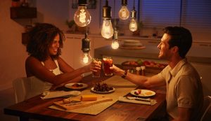 Two people share drinks at a dining table under warm LED pendant lights, with food and fruit displayed in a modern kitchen setting.
