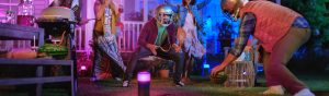 Group of people in a backyard at night, playing with watermelons and wearing football helmets, colorful smart lights illuminate the scene.