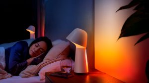 Person lying in bed at night, bedside table lamp casting soft orange light, glass of water and phone on wooden nightstand.