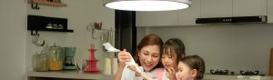 A group in a modern kitchen decorates pastries under a large circular pendant light, with shelves and kitchenware in the background.