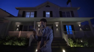 Person standing in front of a house at night using a smartphone, with outdoor smart security lighting illuminating the entrance.