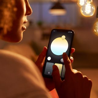 Person adjusting smart lighting color and brightness using a mobile app in a warmly lit indoor setting with visible hanging bulbs.