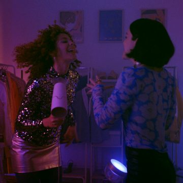 Two people in a room with colorful gradient lighting, one holding a hairdryer, engaging in conversation near clothing racks.
