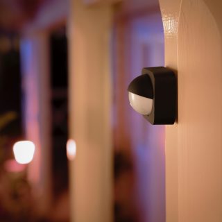 Black and white motion sensor mounted on an exterior wall, positioned to monitor entryway with outdoor smart lights in the background.