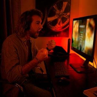 Person celebrating a gaming win at a desk with ambient colored lighting behind a monitor in a home office setting.