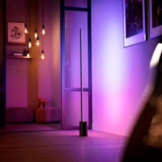 Slim vertical floor lamp placed near a wall, emitting colored light onto the surface, with pendant bulbs in the background.