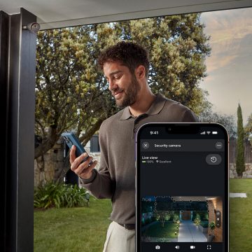 A person in a garden uses a smartphone to monitor home security and lighting through a live view camera app.