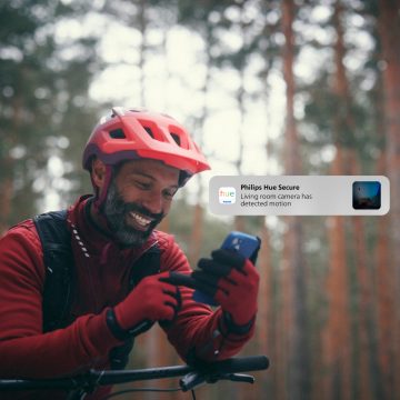 A cyclist in outdoor gear checks a smartphone in a forest, viewing a security notification about detected motion from a living room camera.