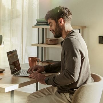 Person working at a desk with a laptop in a softly lit home office, warm light illuminating workspace and shelves in the background.