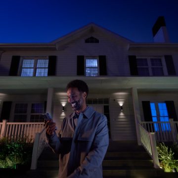 Person standing outside a house at night using a smartphone, with smart outdoor lights illuminating the porch and entryway.