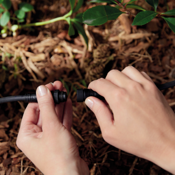 Hands connecting two black outdoor cables, likely for smart outdoor lighting installation, with soil and plants in the background.