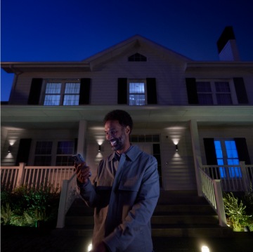 Person standing in front of a house at night using a smartphone, with outdoor smart lighting illuminating the entrance and pathway.