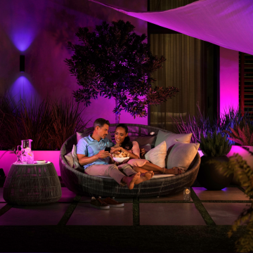 Two people sitting on an outdoor sofa sharing snacks, with purple LED light panels illuminating the garden and wall behind them.