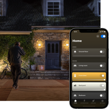 A person approaches a house at night, illuminated by outdoor lights near the front door, with a smart lighting app screen visible.